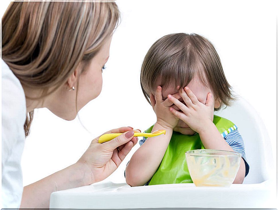 Baby with his hands covering his eyes as his mother offers to eat with a spoon to help him gain weight.